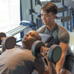 Trainer and man lying and lifting dumbbells in gym