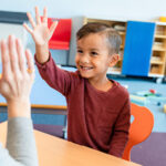 Boy raising hand in classroom with mom and teacher