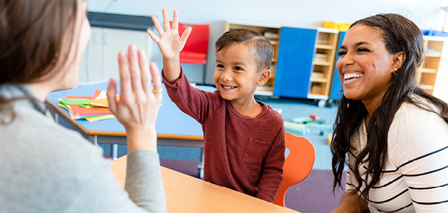 Boy raising hand in classroom with mom and teacher
