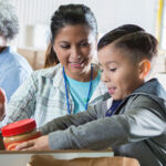 Mid adult mom and her elementary age son sort food items received during a food drive. The little boy is placing a jar of peanut butter into a donation box.