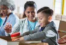 Mid adult mom and her elementary age son sort food items received during a food drive. The little boy is placing a jar of peanut butter into a donation box.