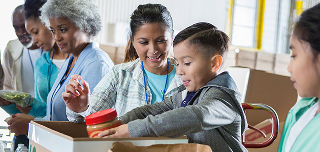 Mid adult mom and her elementary age son sort food items received during a food drive. The little boy is placing a jar of peanut butter into a donation box.